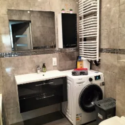 Szara łazienka z umywalką nablatową, czarnymi szafkami pod umywalką, pralką Samsung, grzejnikiem drabinkowym i lustrem w ramie z mozaiki.