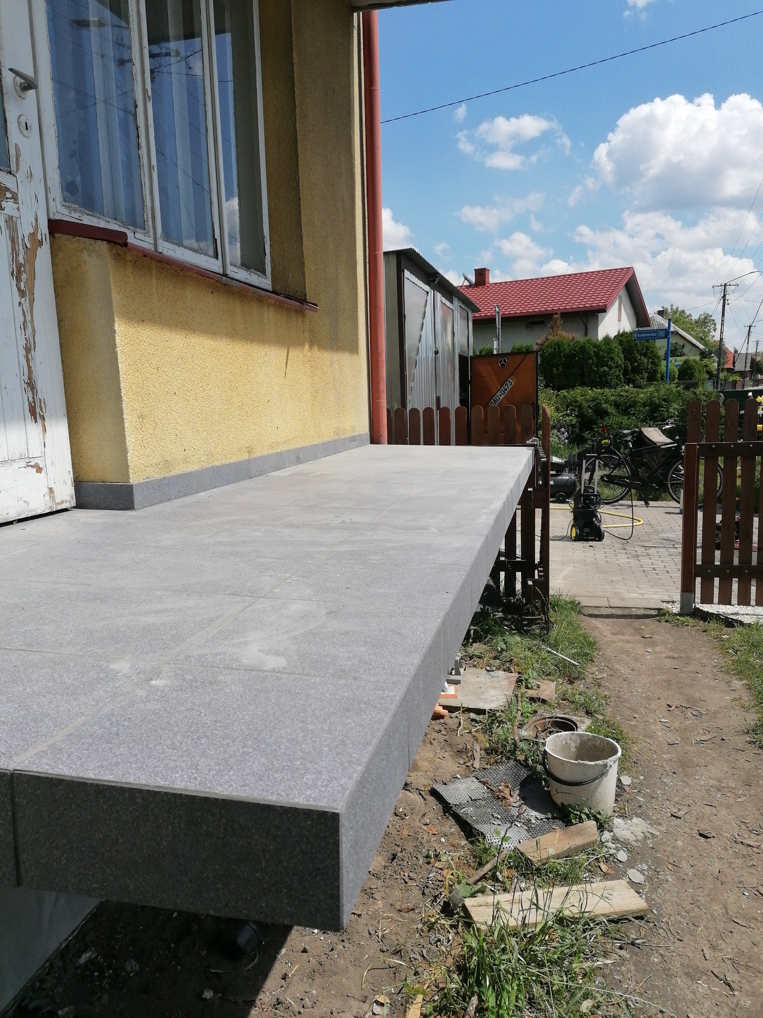 Wykończony, szary, kamienny taras przed domem z żółtą elewacją. Widoczna biała, obdrapana rama okienna i drzwi. W tle rower, myjka ciśnieniowa i drewniane ogrodzenie.