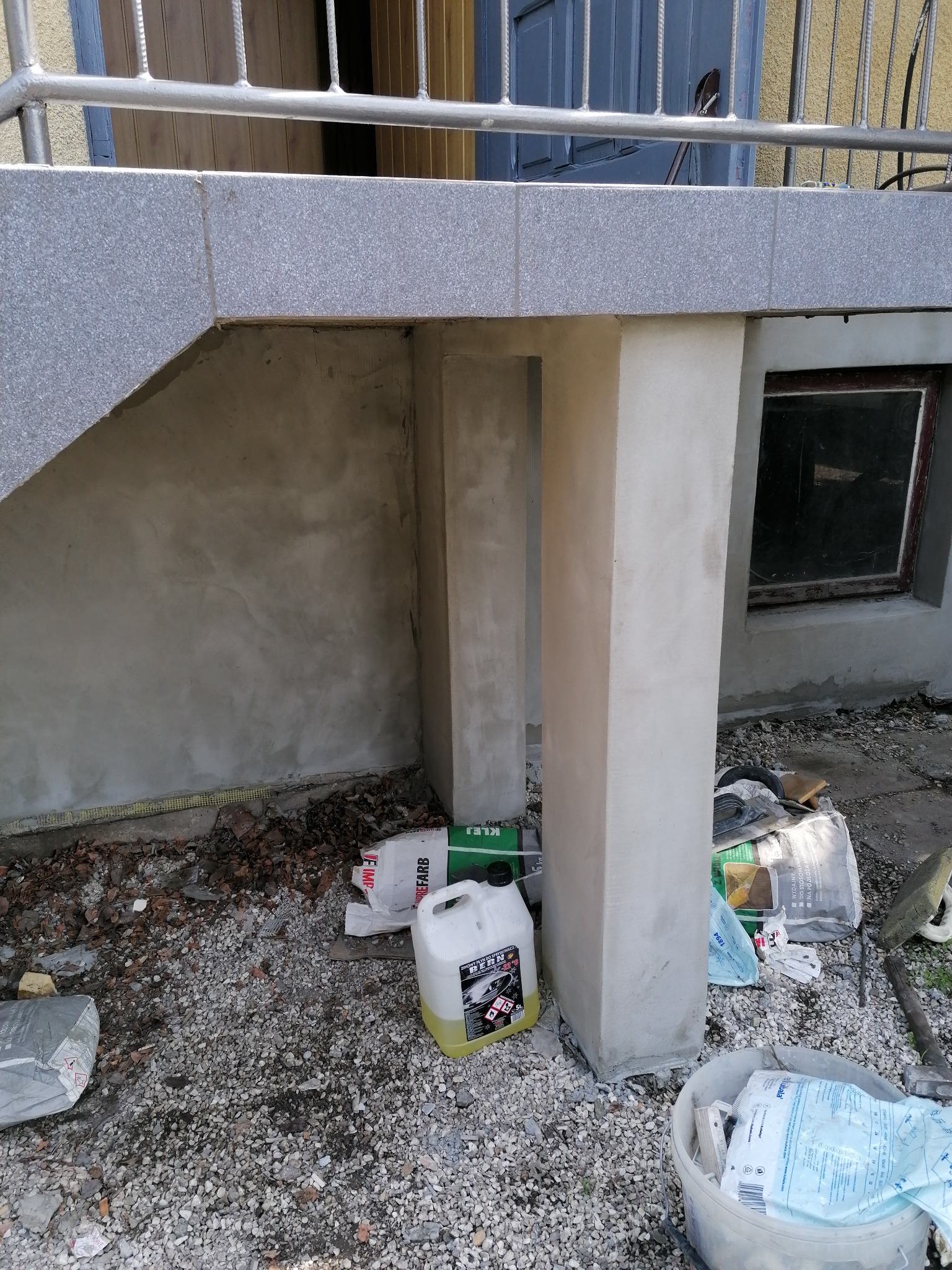 Wykończona podstawa schodów zewnętrznych z otynkowanymi filarami, widoczne materiały budowlane i narzędzia na żwirowym podłożu.