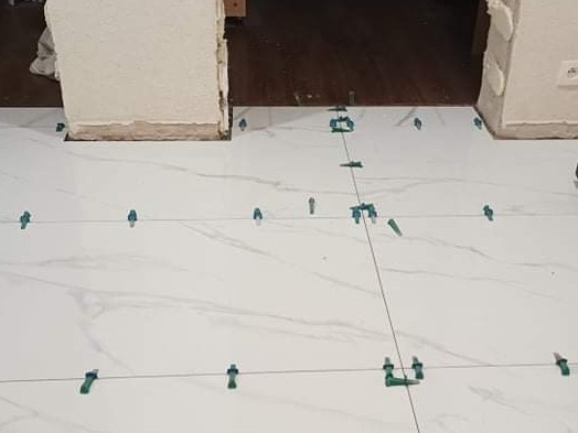 Montaż białych płytek podłogowych z efektem marmuru, z użyciem sznurka i zielonych krzyżyków dystansowych.