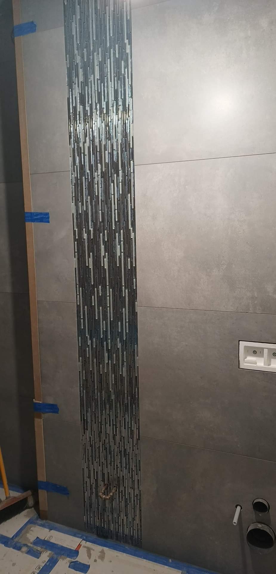 Układanie dekoracyjnej mozaiki z podłużnych płytek w pasie na szarej ścianie w łazience, widoczne taśmy zabezpieczające i elementy instalacji hydraulicznej.