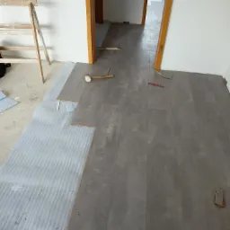 Proces układania szarych paneli podłogowych w korytarzu, widoczna pianka podkładowa, młotek, ołówek i drewniana drabina oparta o ścianę.