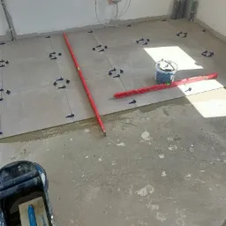 Układanie szarych płytek podłogowych z użyciem poziomicy i systemu poziomowania, widoczne narzędzia glazurnicze i instalacje na ścianie.