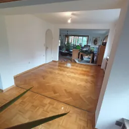 Jasny parkiet ułożony w jodełkę w przejściu między pokojami, świeżo pomalowane białe ściany, widok na salon z roślinami.