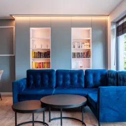 Niebieski, welurowy narożnik z pikowanymi oparciami i okrągłymi stolikami kawowymi na drewnianej podłodze, w tle zabudowa ściany z półkami na książki i dekoracje.