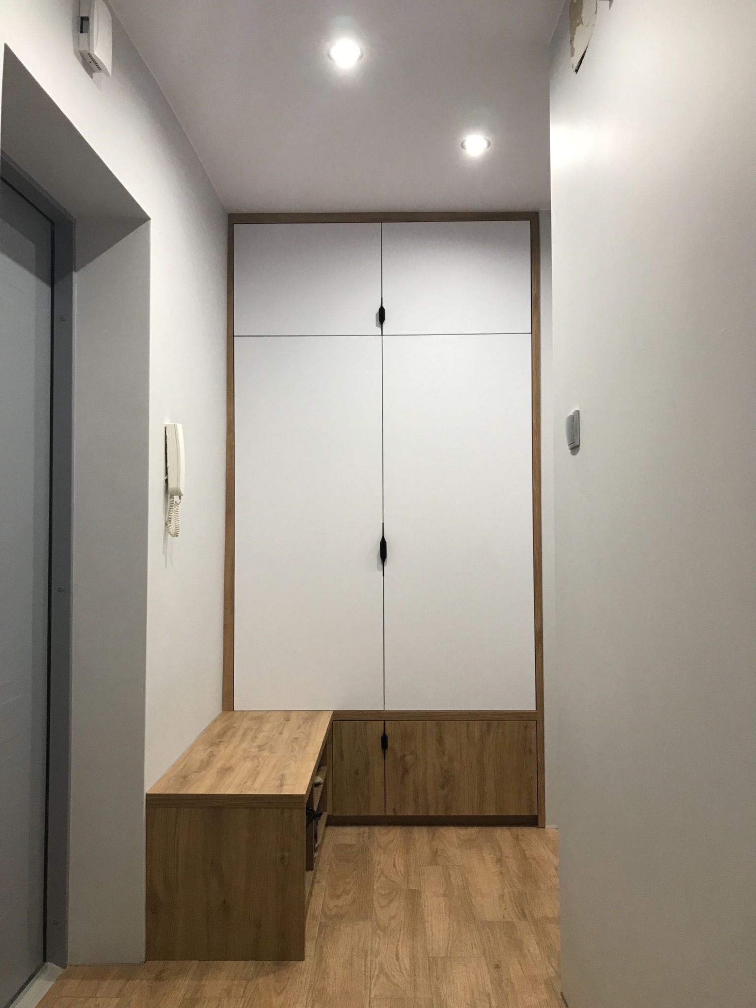 Biała szafa w zabudowie z drewnianą ramą i siedziskiem w przedpokoju, czarne podłużne uchwyty, drewniana podłoga.