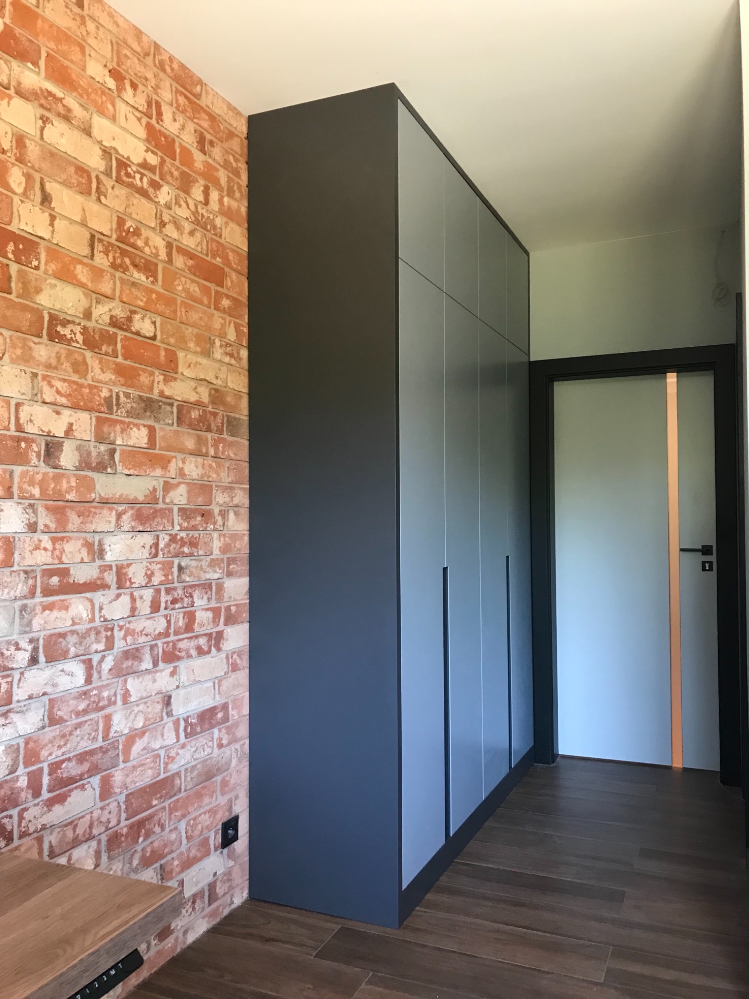 Wysoka, grafitowa zabudowa meblowa z szarymi frontami, dopasowana do wnęki, na tle ściany z czerwonej cegły i brązowej podłogi imitującej drewno.