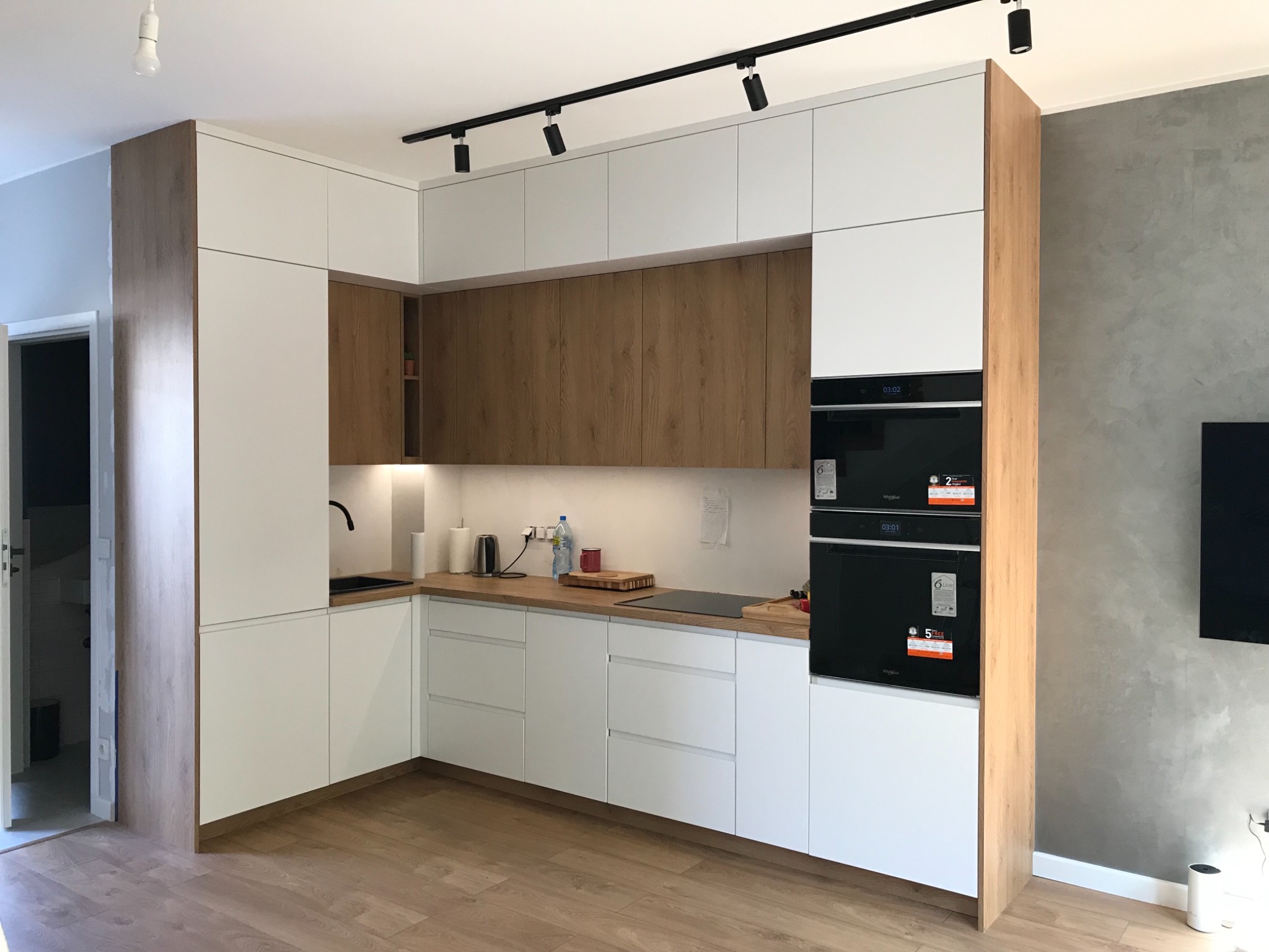 Nowoczesna kuchnia z białymi frontami i drewnianymi akcentami, oświetlenie szynowe, sprzęt AGD w zabudowie, widok na drzwi do innego pomieszczenia.
