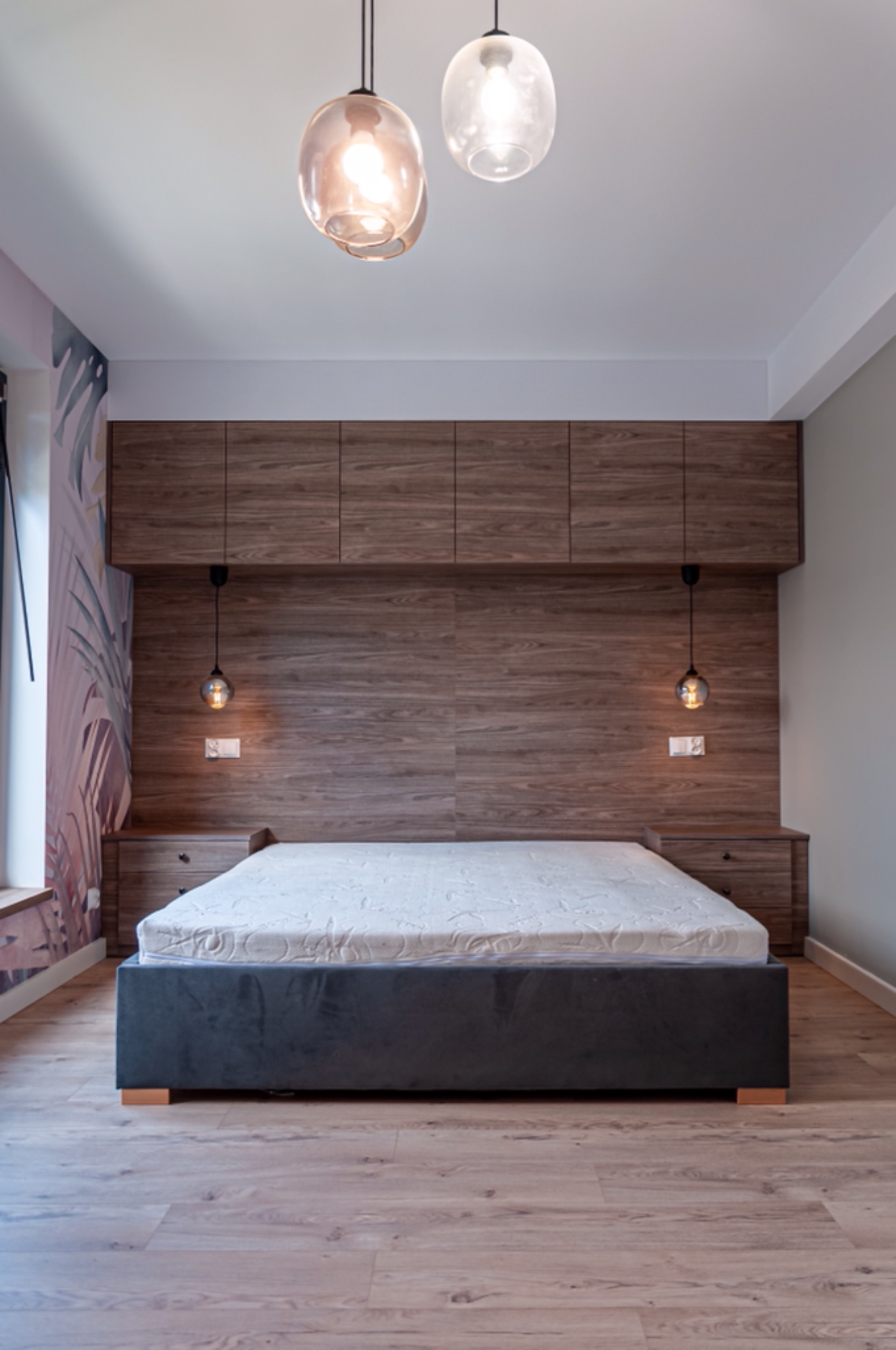 Sypialnia z łóżkiem kontynentalnym na drewnianej podłodze, z wezgłowiem z paneli drewnopodobnych z szafkami nocnymi i szafkami wiszącymi, oraz lampami wiszącymi z okrągłymi kloszami.