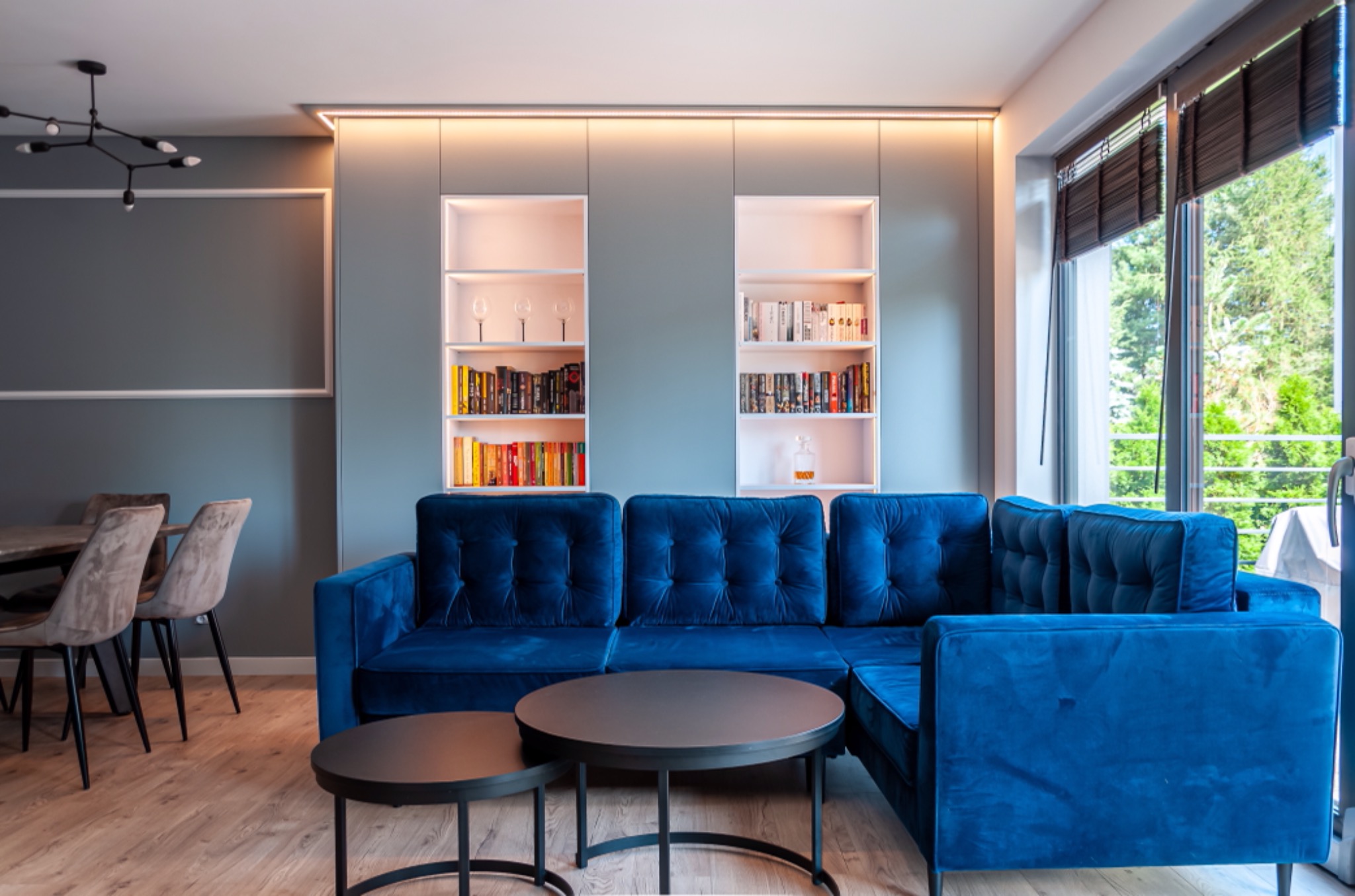 Niebieski, welurowy narożnik z pikowanymi oparciami i okrągłymi stolikami kawowymi na drewnianej podłodze, w tle zabudowa ściany z półkami na książki i dekoracje.