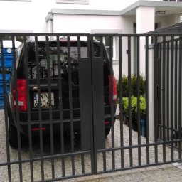 Astern - Nowoczesna, czarna, metalowa brama dwuskrzydłowa z pionowymi prętami, zamontowana na białych, murowanych słupkach, widok z zewnątrz na podjazd z kostki brukowej i zaparkowany czarny samochód.