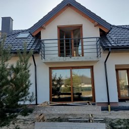 Astern - Nowoczesny dom jednorodzinny w trakcie budowy z widoczną czarną, metalową balustradą balkonową, drewnianą stolarką okienną i czarnym dachem z dachówki ceramicznej.