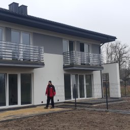 Astern - Nowoczesny, dwukondygnacyjny dom w trakcie wykończenia, z balkonami z barierkami zabezpieczonymi folią, widoczny pracownik w czerwonej kurtce i ogrodzenie panelowe.