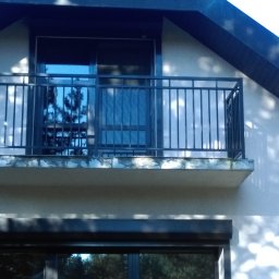 Astern - Czarna, metalowa balustrada balkonowa z pionowymi szczeblami, widoczna z zewnątrz budynku, z fragmentem zwiniętej markizy po lewej stronie.