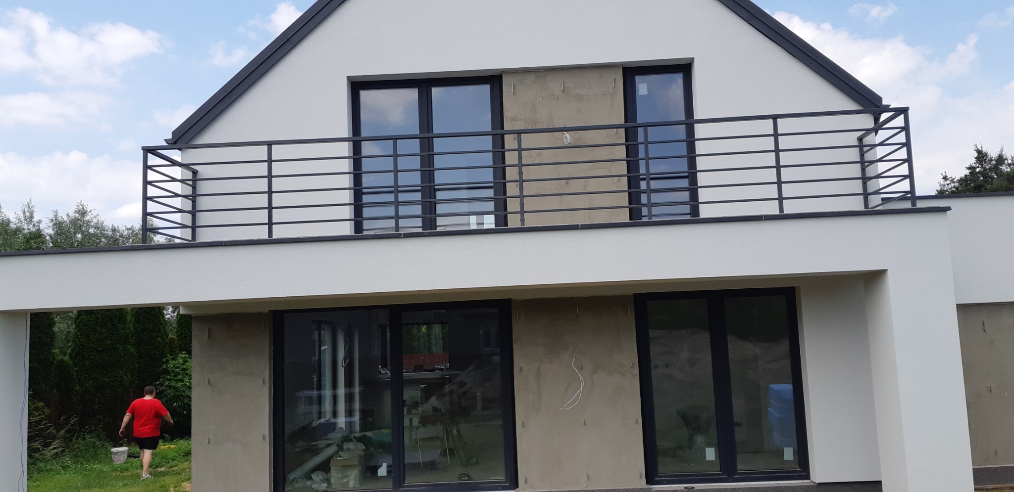 Nowoczesny dom z balkonem zabezpieczonym czarną, metalową balustradą. Widoczne okna z czarnymi ramami i fragment ogrodu z zielenią. Ściany domu częściowo w stanie surowym.