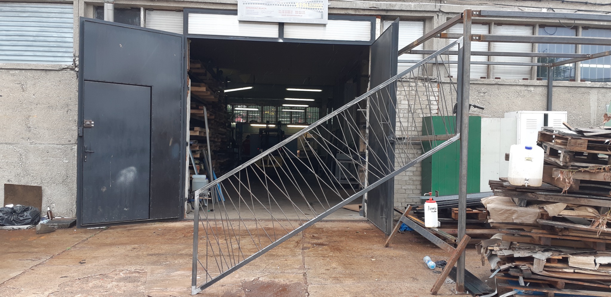 Metalowa konstrukcja schodów z prętów, w trakcie produkcji, widoczna na tle otwartej hali warsztatowej z materiałami i narzędziami.