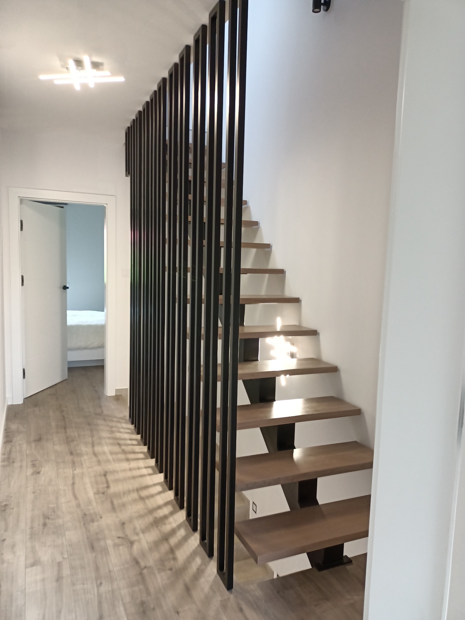 Nowoczesne schody drewniane z czarnymi metalowymi wspornikami i pionowymi elementami zabezpieczającymi, widok z dołu, minimalistyczne wnętrze z otwartymi drzwiami do sypialni w tle.