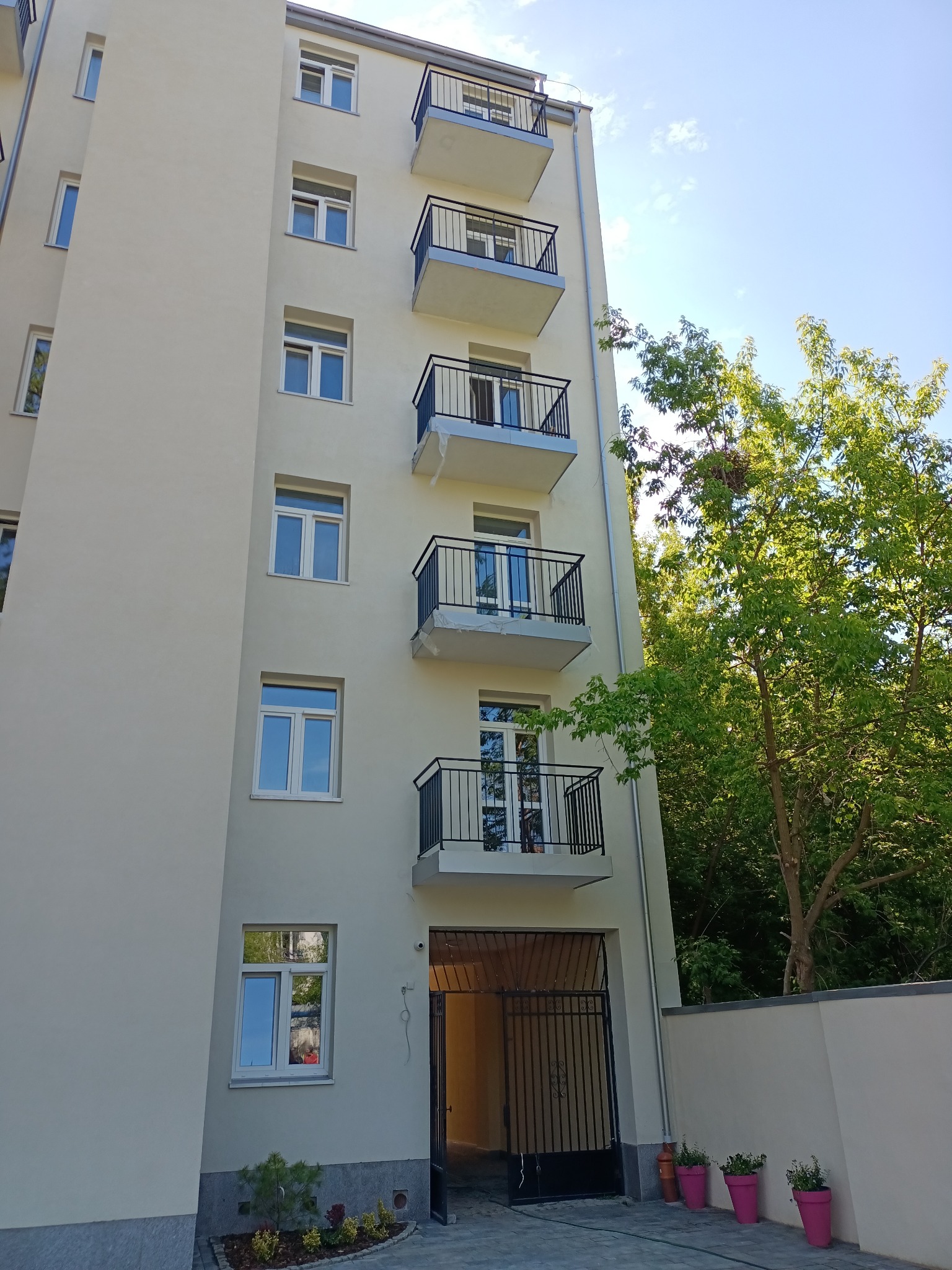 Elewacja budynku wielorodzinnego z balkonami z czarnymi, metalowymi balustradami, widok z dołu na tle zieleni i błękitnego nieba.