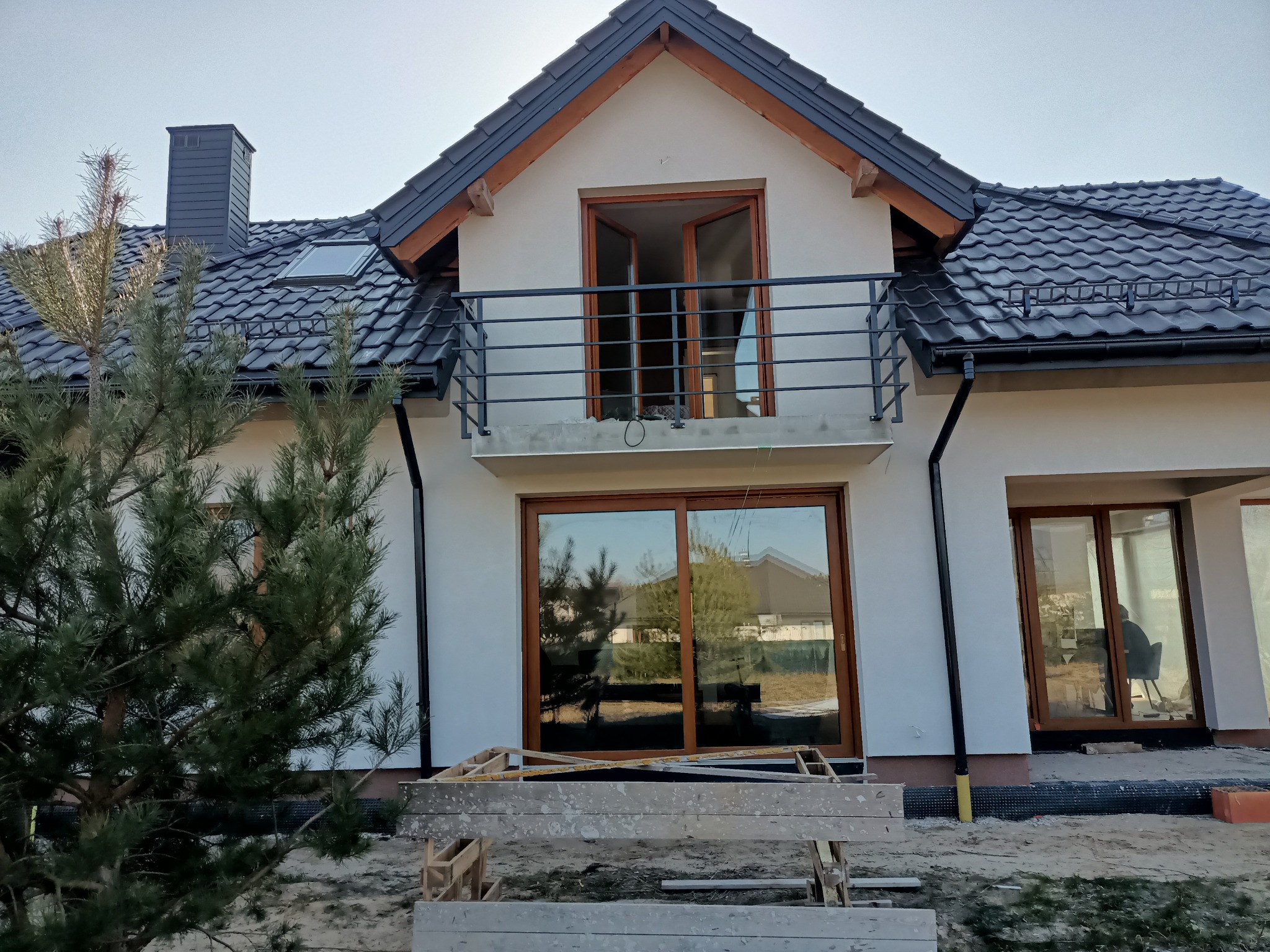 Nowoczesny dom jednorodzinny w trakcie budowy z widoczną czarną, metalową balustradą balkonową, drewnianą stolarką okienną i czarnym dachem z dachówki ceramicznej.