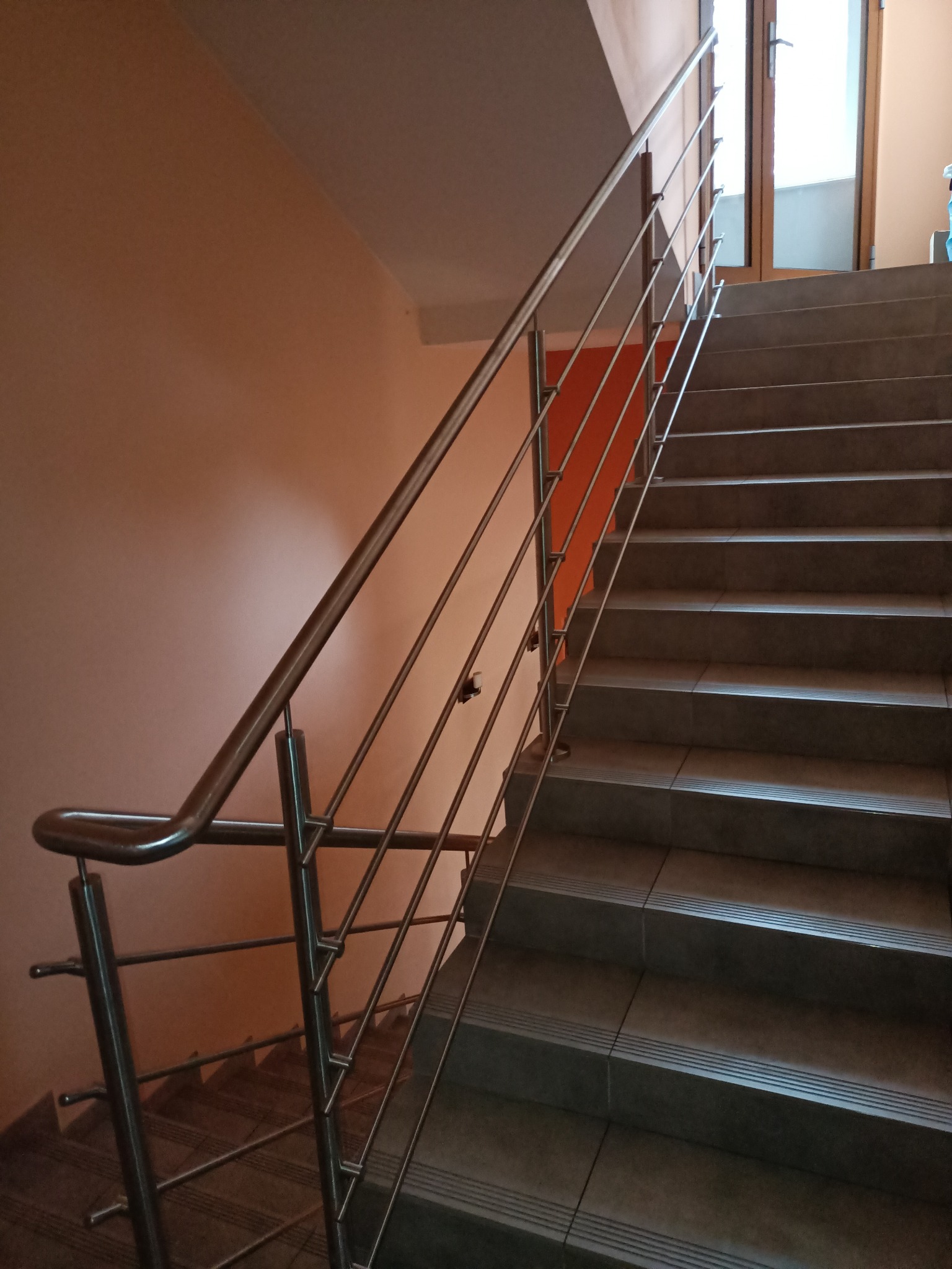 Klatka schodowa z szarymi płytkami i prostą balustradą ze stali nierdzewnej z pionowymi prętami, widok z dołu.