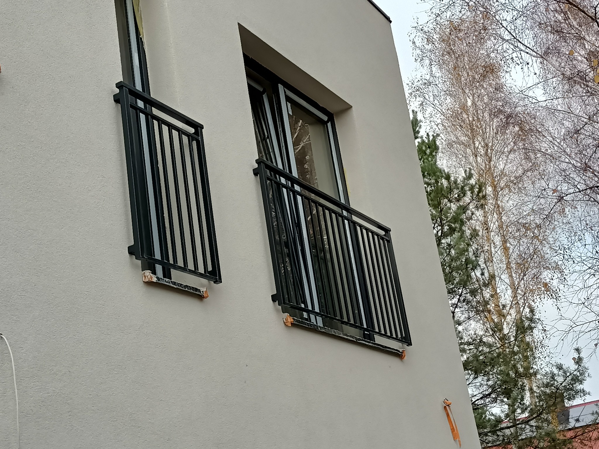 Nowoczesna, czarna balustrada balkonowa z metalowymi prętami, zamontowana na elewacji budynku w trakcie wykańczania, z widocznymi elementami montażowymi i drzewami w tle.