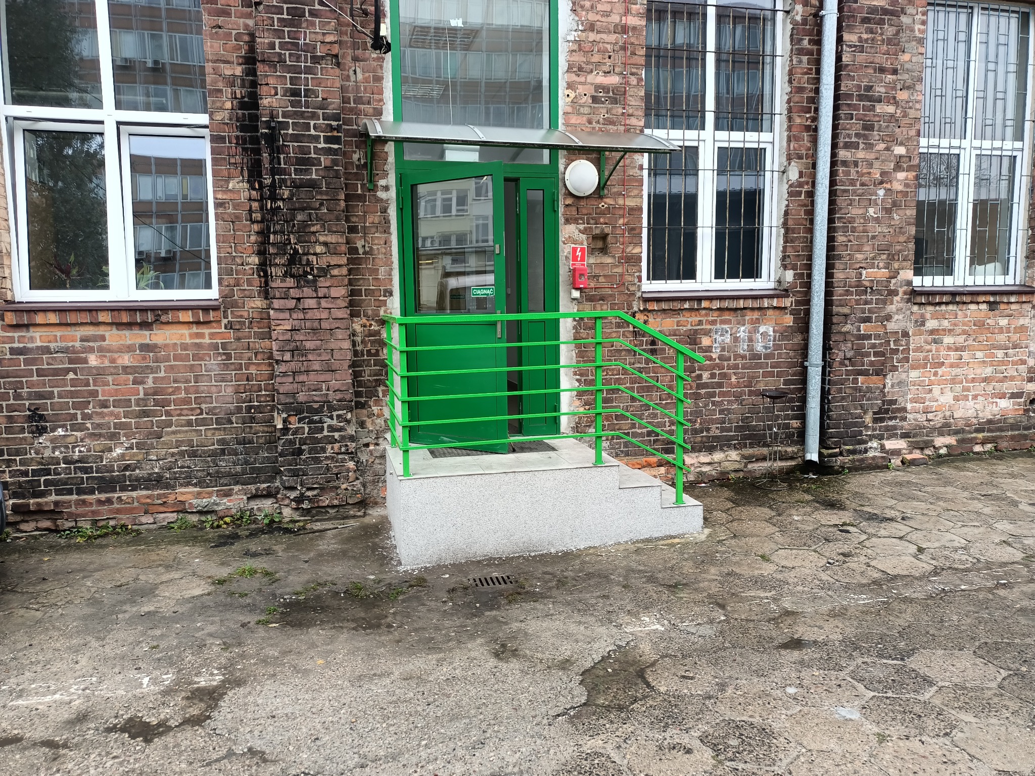 Zewnętrzne wejście do budynku z cegły z zielonymi ramami okiennymi i zieloną balustradą z poręczą. Widoczne dwa stopnie prowadzące do drzwi.