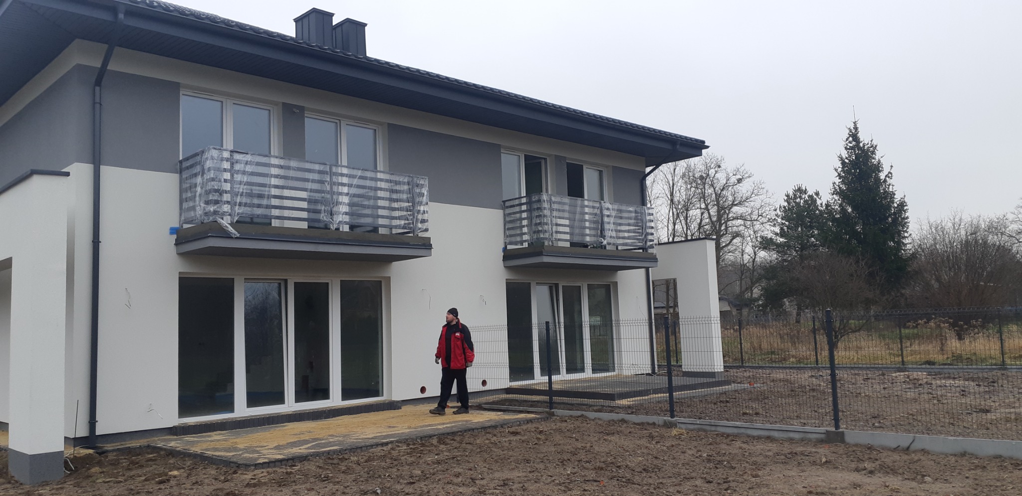 Nowoczesny, dwukondygnacyjny dom w trakcie wykończenia, z balkonami z barierkami zabezpieczonymi folią, widoczny pracownik w czerwonej kurtce i ogrodzenie panelowe.
