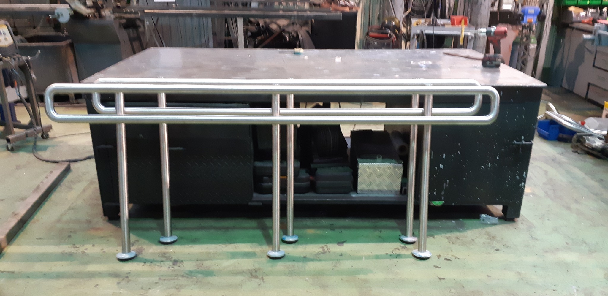 Nierdzewna poręcz z zaokrąglonymi elementami, ustawiona na warsztatowym stole, widoczne narzędzia i elementy obróbki metalu w tle.