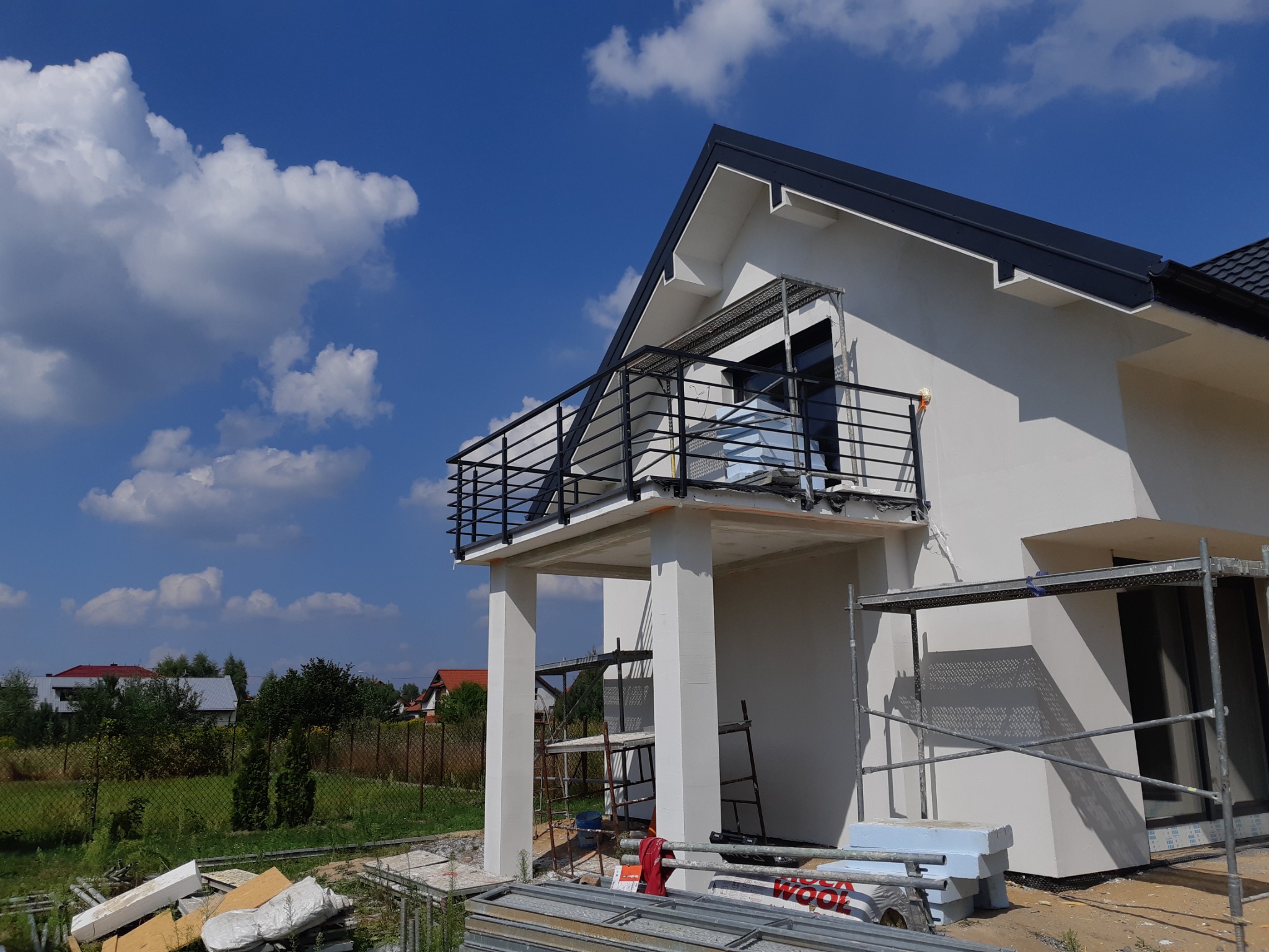 Nowoczesny dom jednorodzinny z balkonem zabezpieczonym czarną, metalową balustradą, widoczne rusztowania i materiały budowlane.