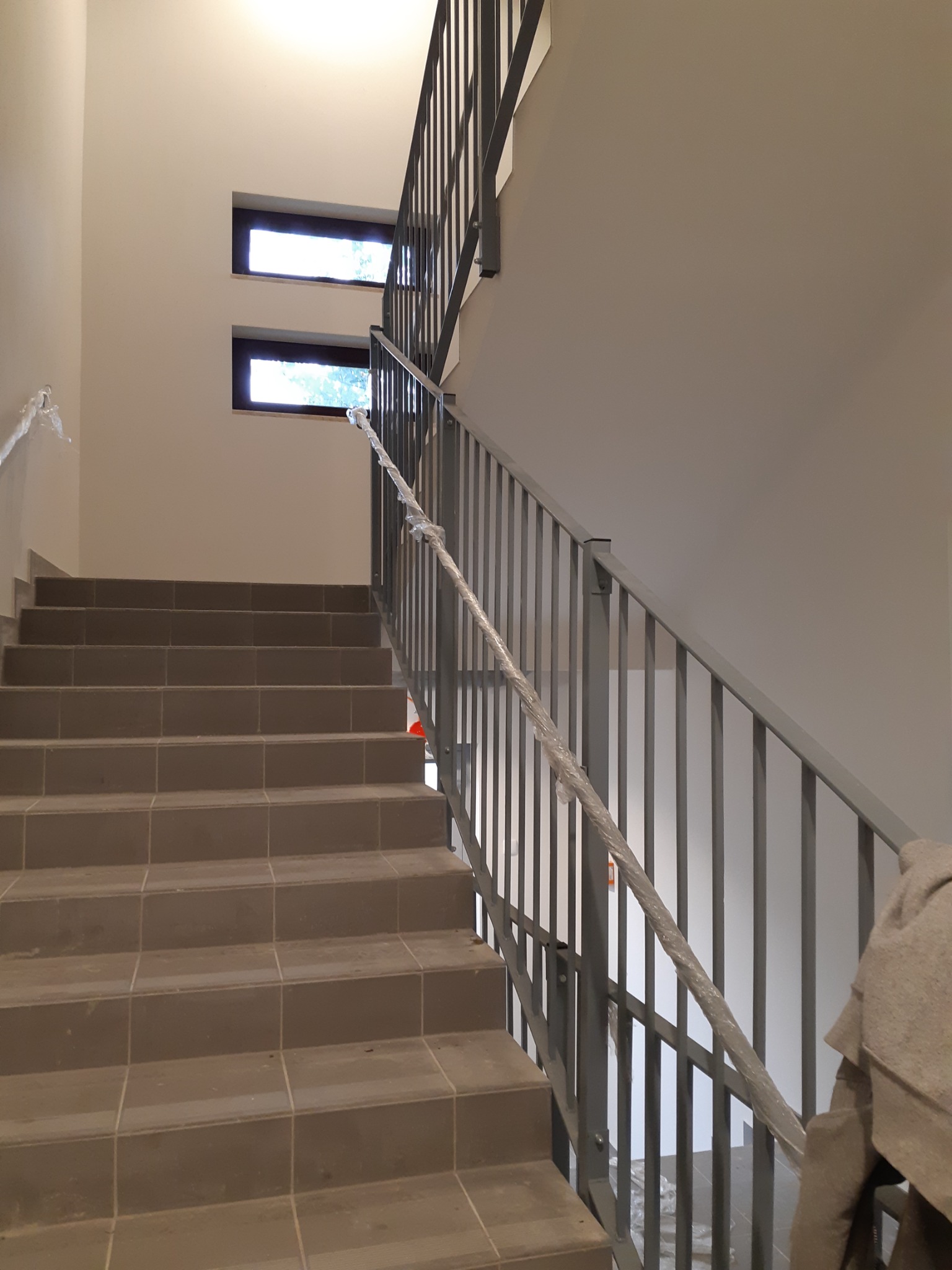 Nowoczesne schody z szarymi stopniami i metalową balustradą, zabezpieczoną folią ochronną podczas prac wykończeniowych, widoczne dwa okna w tle.