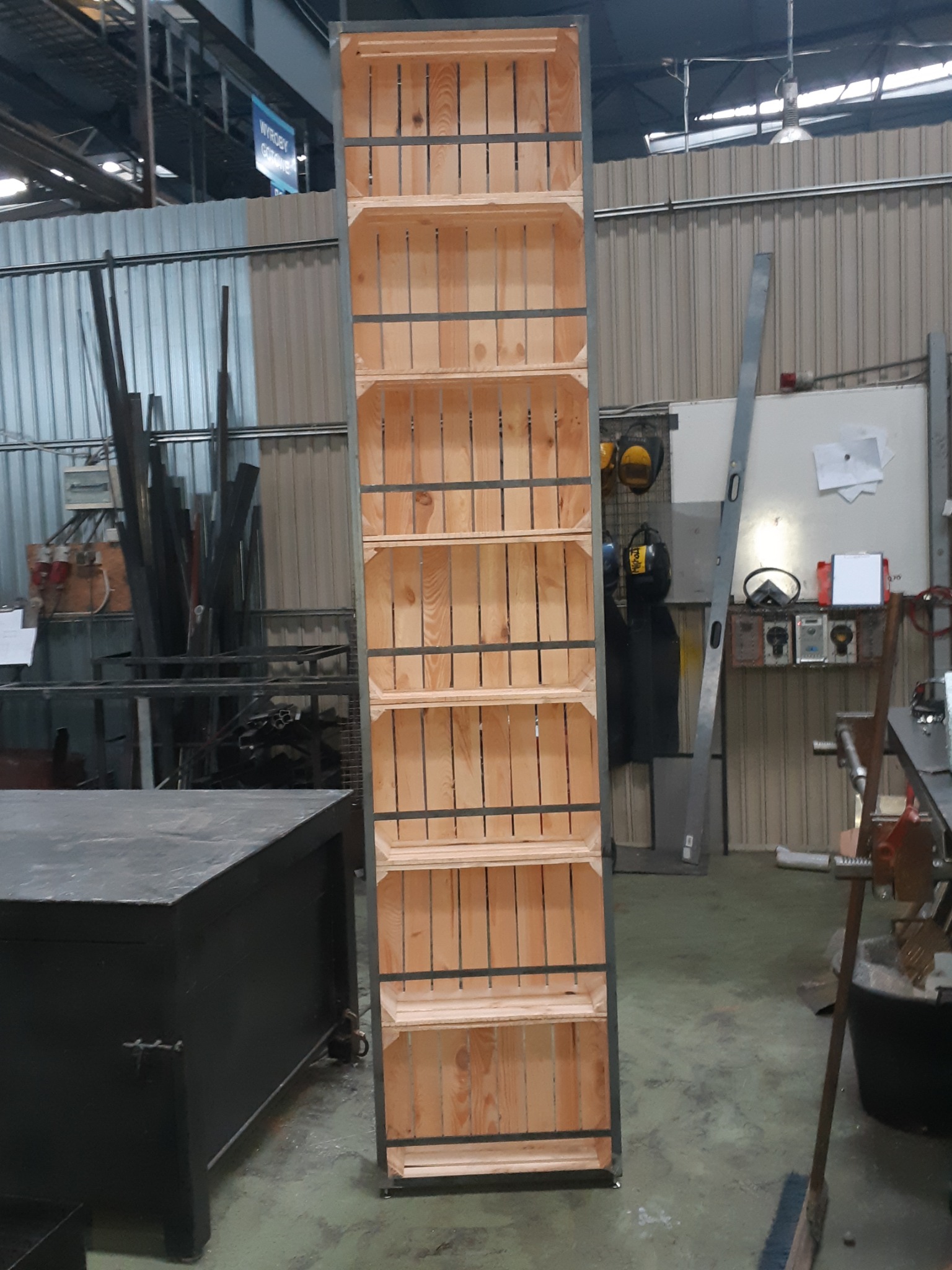 Wysoki regał z drewnianych skrzynek z metalową ramą, wykonany w warsztacie w Warszawie, widoczny na tle narzędzi i elementów konstrukcyjnych.