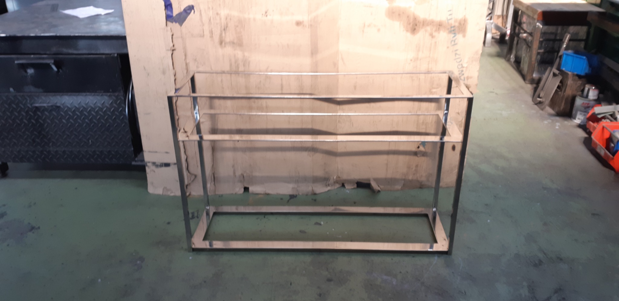 Metalowa konstrukcja regału z profili zamkniętych, spawana, w surowym stanie, na tle kartonów i narzędzi w warsztacie.