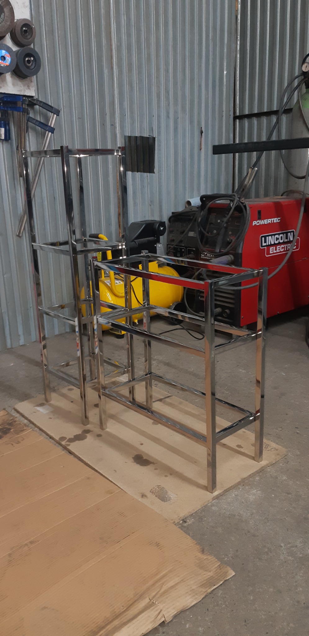 Dwie metalowe konstrukcje szkieletowe z rur, prawdopodobnie w trakcie produkcji w warsztacie, z widocznym sprzętem spawalniczym w tle.