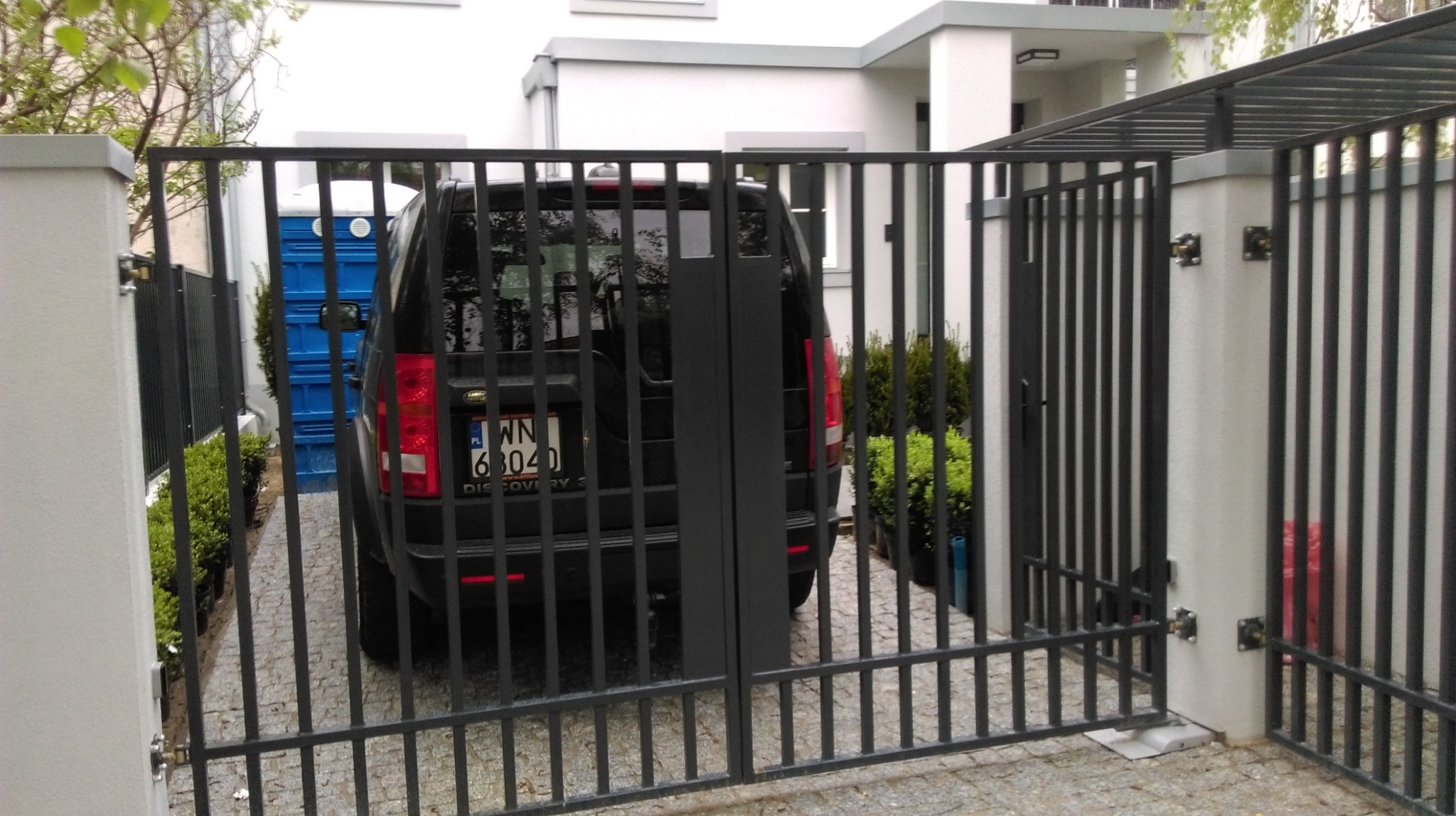 Czarna metalowa brama wjazdowa z pionowymi prętami, widok od strony ulicy na zaparkowany samochód na posesji, w tle biały budynek i niebieska toaleta przenośna.