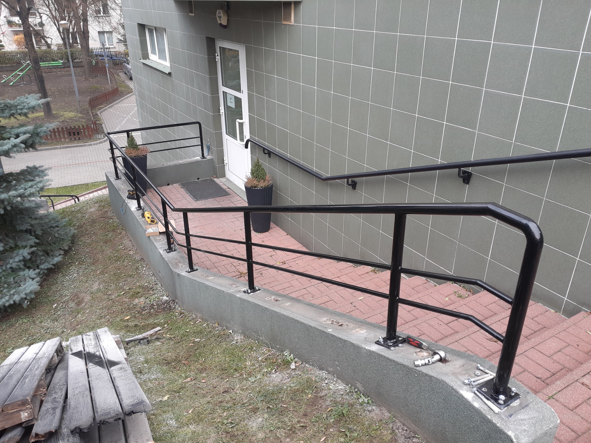 Czarne metalowe balustrady zamontowane na schodach wejściowych do budynku z szarymi płytkami, widoczne narzędzia montażowe i drewniane deski obok.