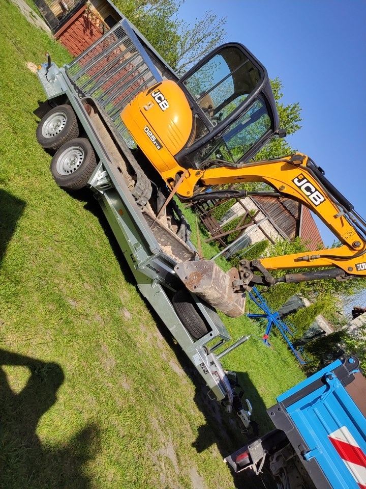 Żółta mini koparka JCB na przyczepie transportowej w Pińczowie, gotowa do transportu na plac budowy, widok z boku na tle zielonego trawnika i zabudowań.