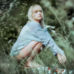 Zamyślona blondynka w ażurowym swetrze kuca na pniu w otoczeniu traw i zieleni, spoglądając w lewo. Ujęcie w plenerze z naturalnym oświetleniem.