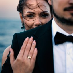 Dawid Ladach - Portret pary: kobieta zza ramienia mężczyzny w smokingu, w tle woda i niebo o zachodzie słońca. Wiatr rozwiewa włosy kobiety, która patrzy w obiektyw.