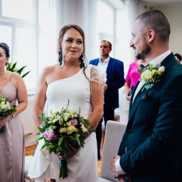 Dawid Ladach - Para młoda podczas ceremonii ślubnej w jasnym wnętrzu, panna młoda w białej sukni z bukietem, pan młody w ciemnozielonym garniturze z butonierką. Goście w tle.