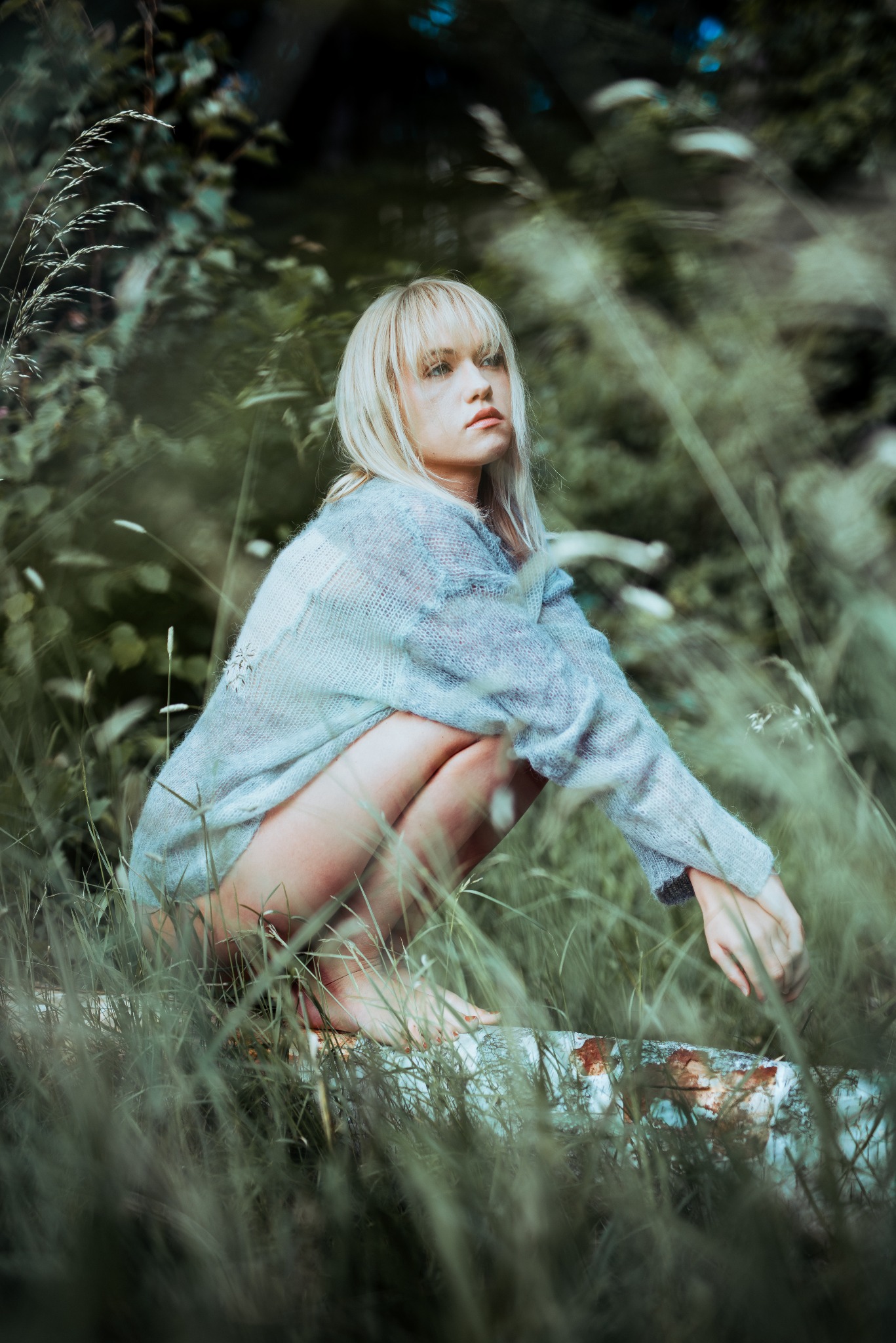 Zamyślona blondynka w ażurowym swetrze kuca na pniu w otoczeniu traw i zieleni, spoglądając w lewo. Ujęcie w plenerze z naturalnym oświetleniem.