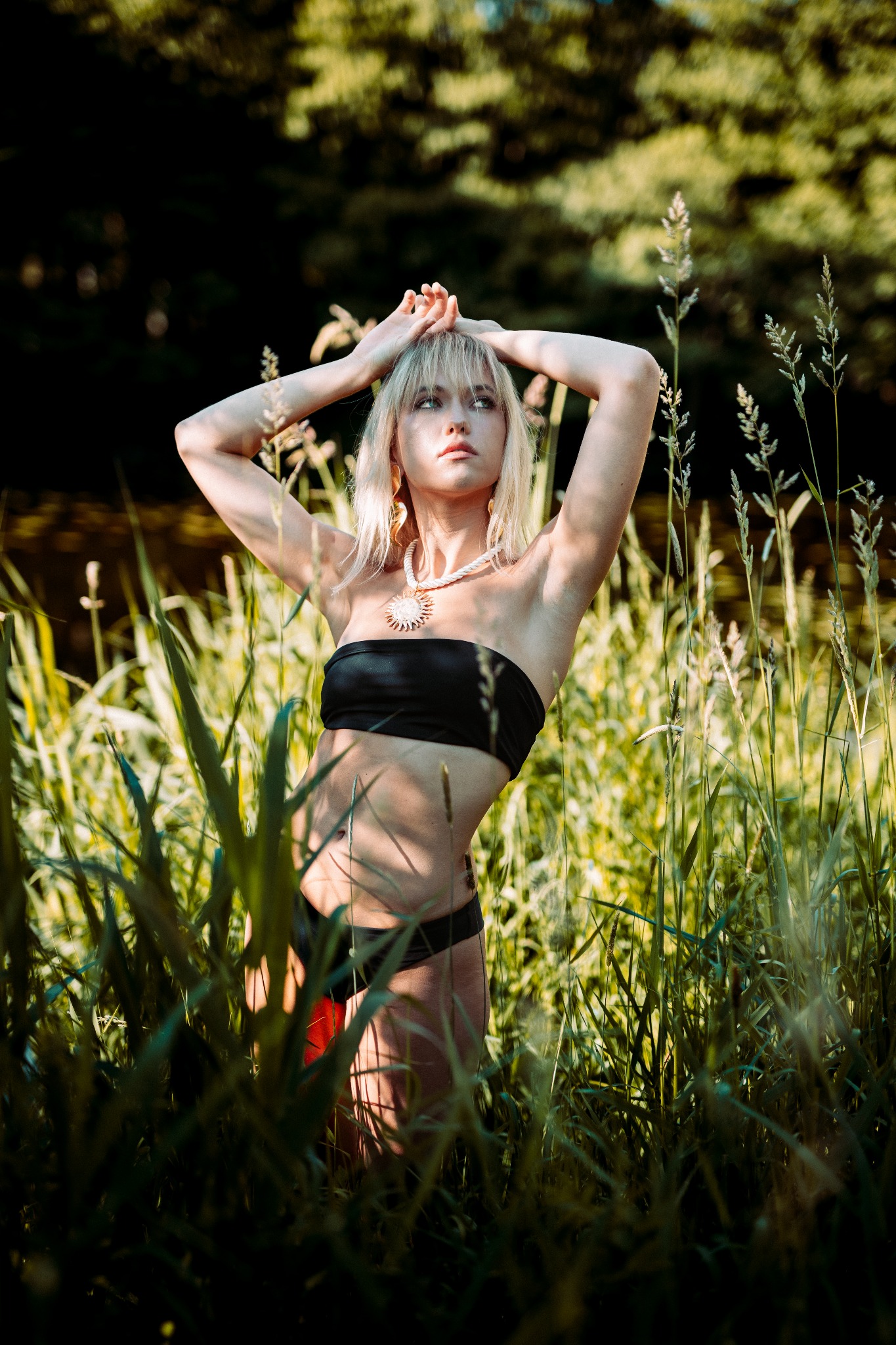 Młoda kobieta w czarnym bikini i ozdobnej biżuterii pozuje w wysokiej trawie, z rękami uniesionymi nad głową. Naturalne światło słoneczne i zielone tło.