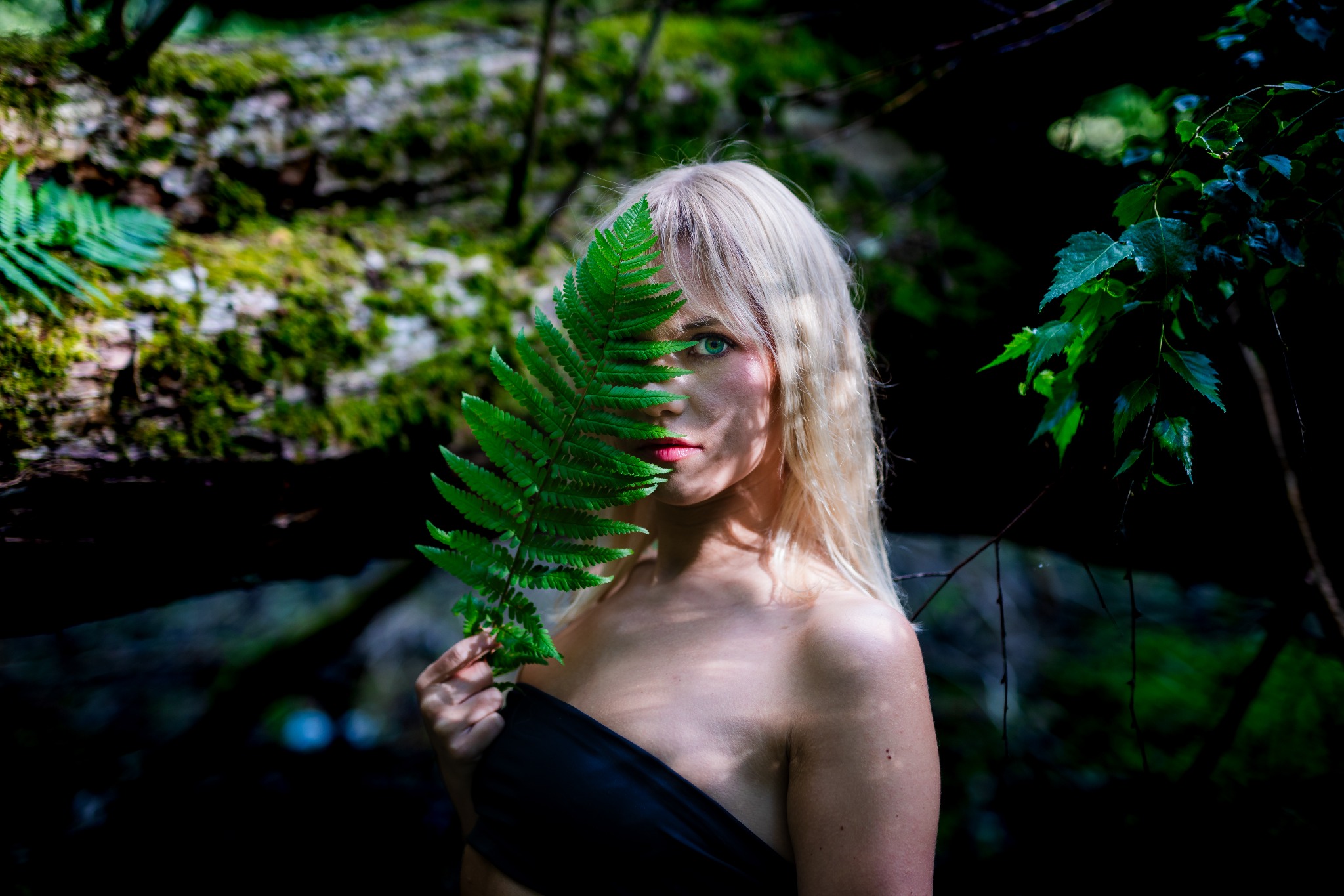 Portret kobiety z blond włosami, zasłaniającej twarz liściem paproci na tle leśnej scenerii w ciemnych tonacjach. Ujęcie z bliska, skupione na spojrzeniu.