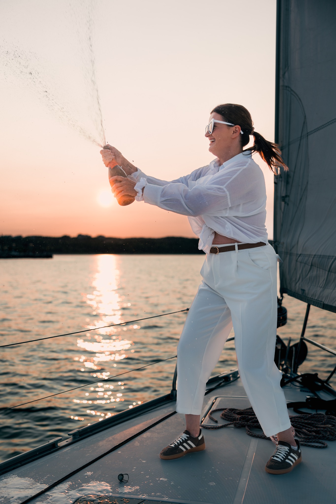 Uśmiechnięta kobieta w białym stroju otwiera szampana na jachcie o zachodzie słońca, w tle woda i zalesiony brzeg. Woda pryska z butelki.