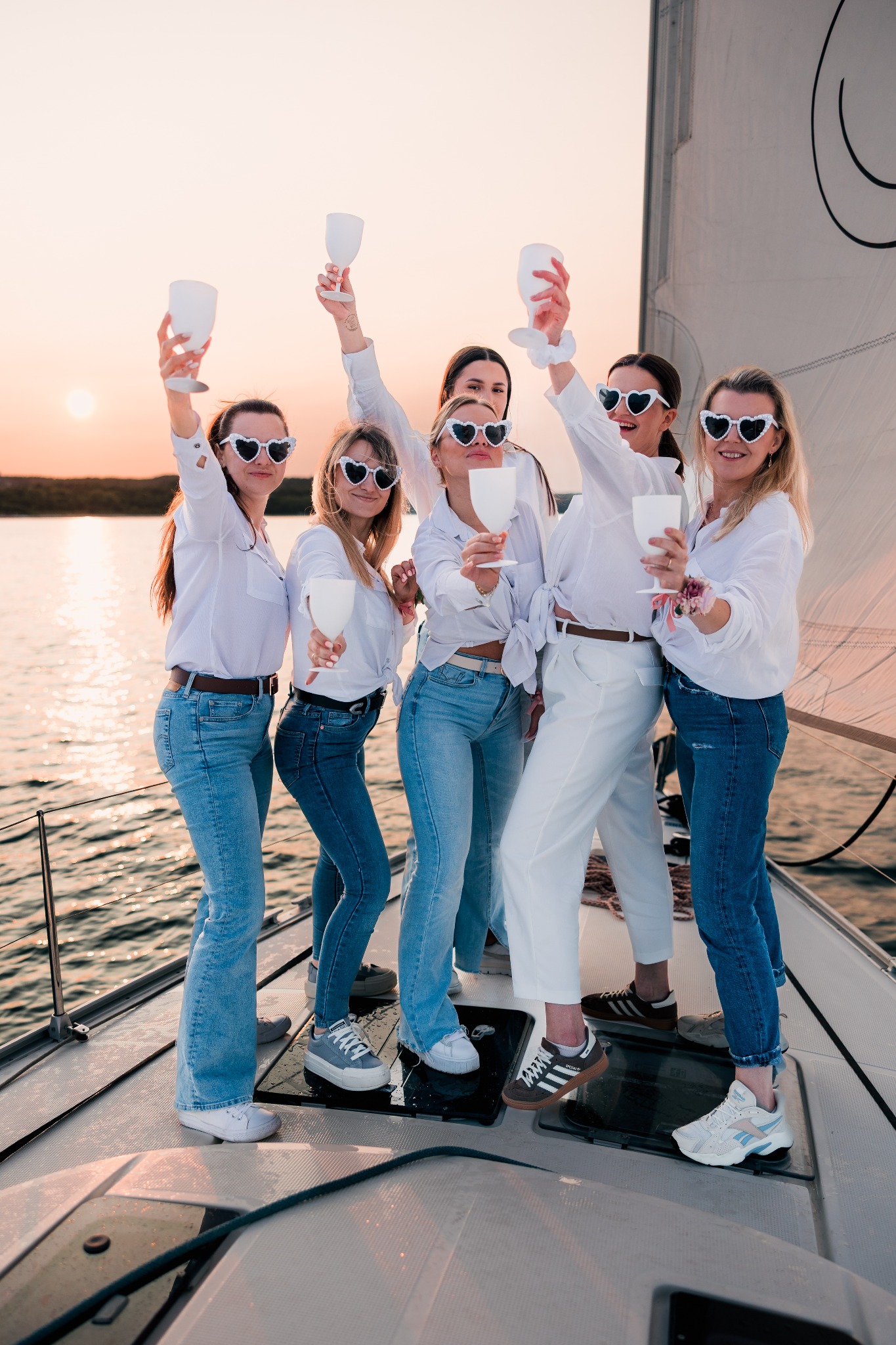 Grupa kobiet w białych koszulach i okularach w kształcie serc wznosi toast białymi kieliszkami na jachcie o zachodzie słońca. Widok na wodę i żagiel.