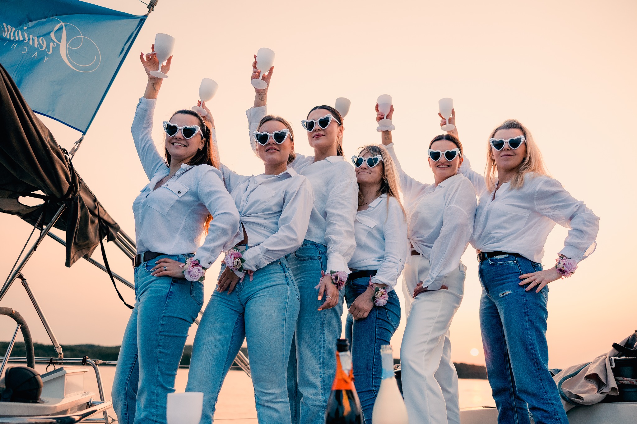 Grupa kobiet w białych koszulach i dżinsach, w okularach w kształcie serc, świętuje z kieliszkami na jachcie o zachodzie słońca. Widoczny maszt i butelki.