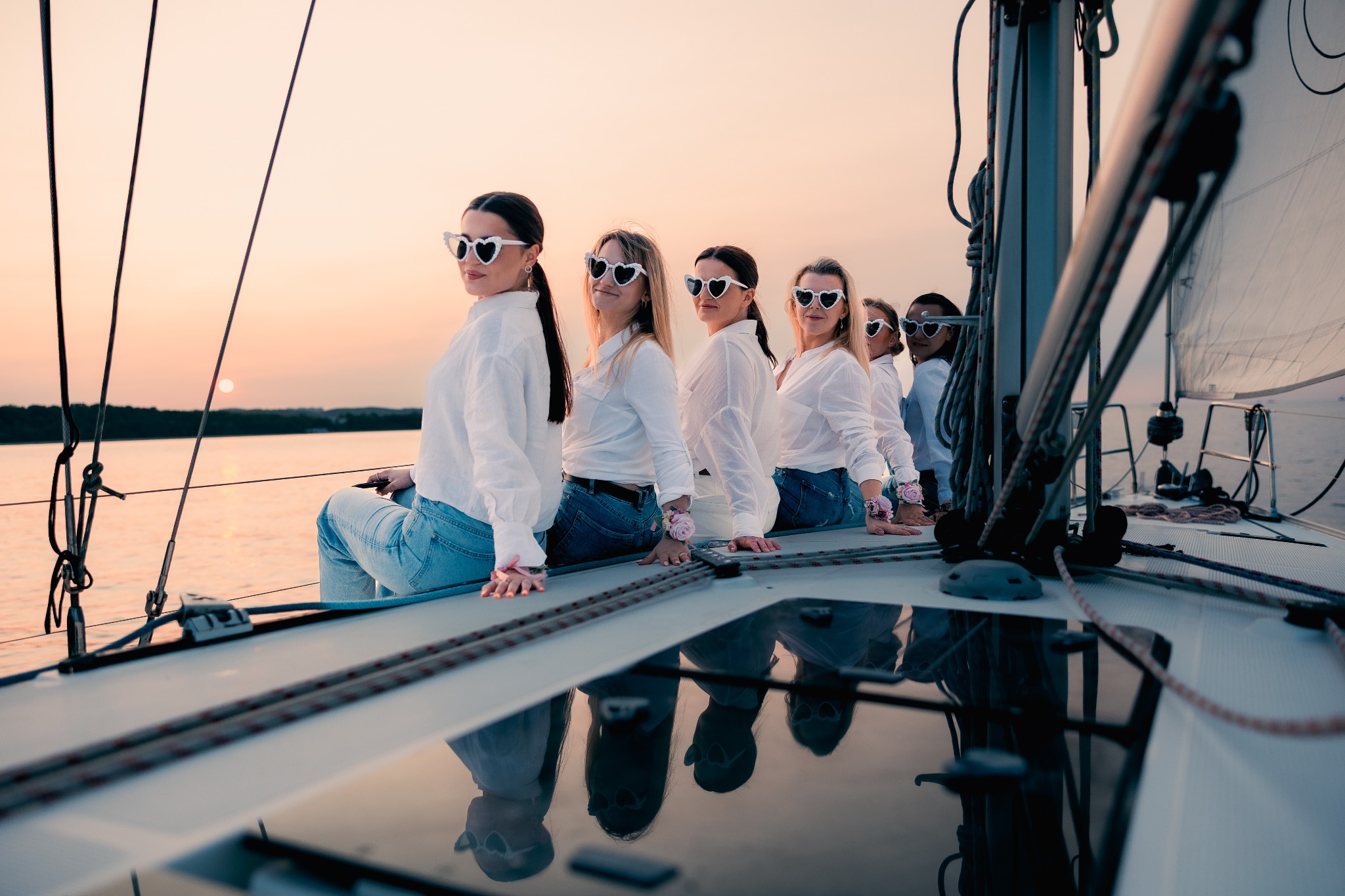Grupa kobiet w białych koszulach i jeansach, z okularami w kształcie serc, siedząca na pokładzie jachtu podczas zachodu słońca. Widoczne liny i maszt.