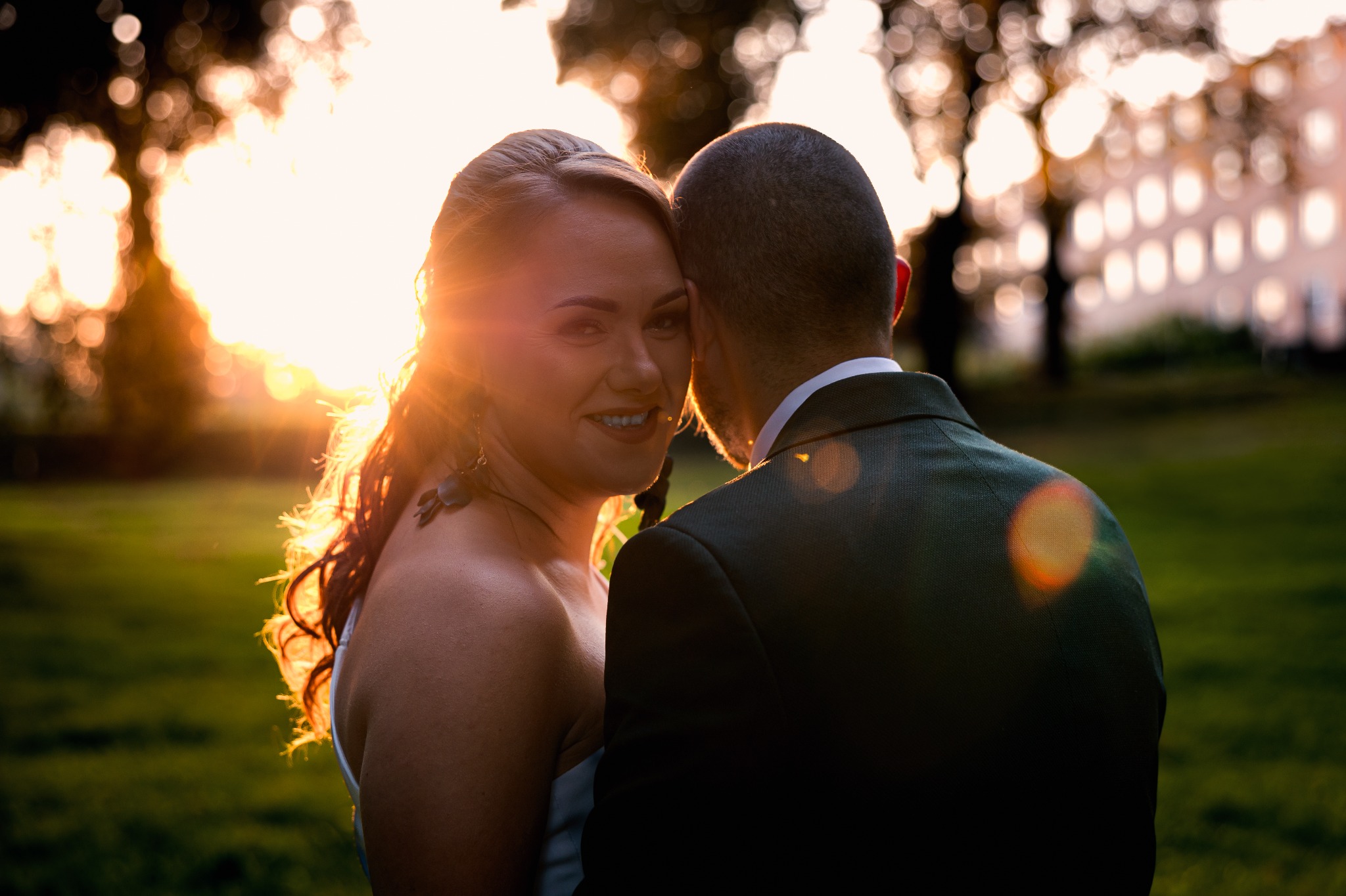 Para młoda w objęciach, panna młoda patrzy w obiektyw, ciepłe światło zachodzącego słońca, zieleń trawy i budynek w tle, ujęcie zza pleców pana młodego.