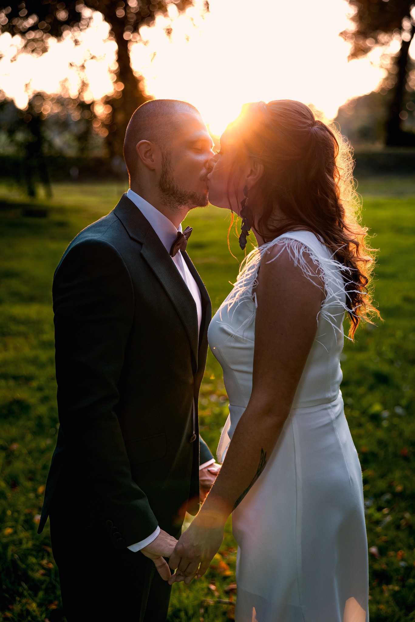 Pocałunek pary młodej w kontraście ze słońcem, ona w białej sukni z piórami, on w ciemnozielonym garniturze z muchą, trzymają się za ręce na tle zieleni.