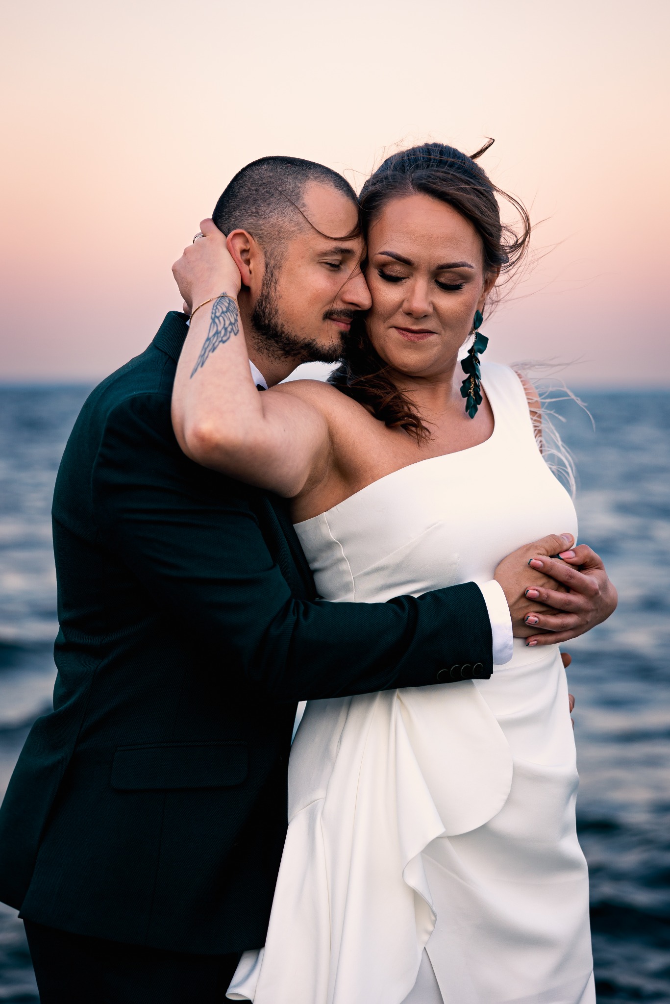 Para młoda w objęciach na tle morza i pastelowego nieba o zachodzie słońca. Pan młody w ciemnozielonym garniturze, panna młoda w białej sukni z asymetrycznym ramieniem.
