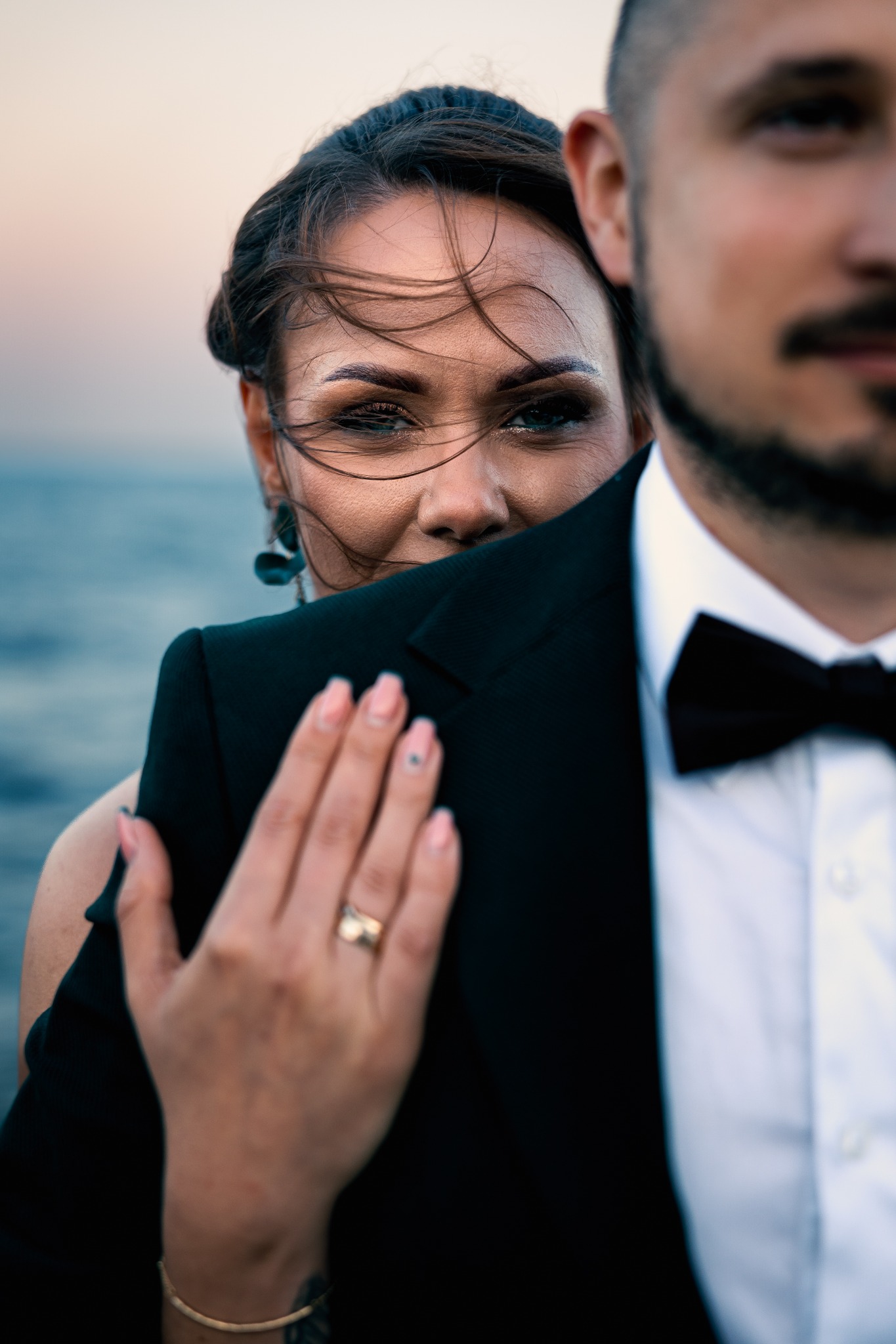 Portret pary: kobieta zza ramienia mężczyzny w smokingu, w tle woda i niebo o zachodzie słońca. Wiatr rozwiewa włosy kobiety, która patrzy w obiektyw.